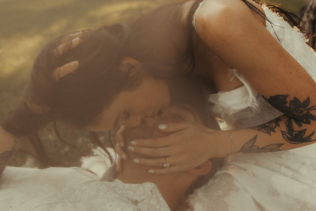 Woman with tattoos on her arms leans down to kiss her fiance as he lays his head in her lap.