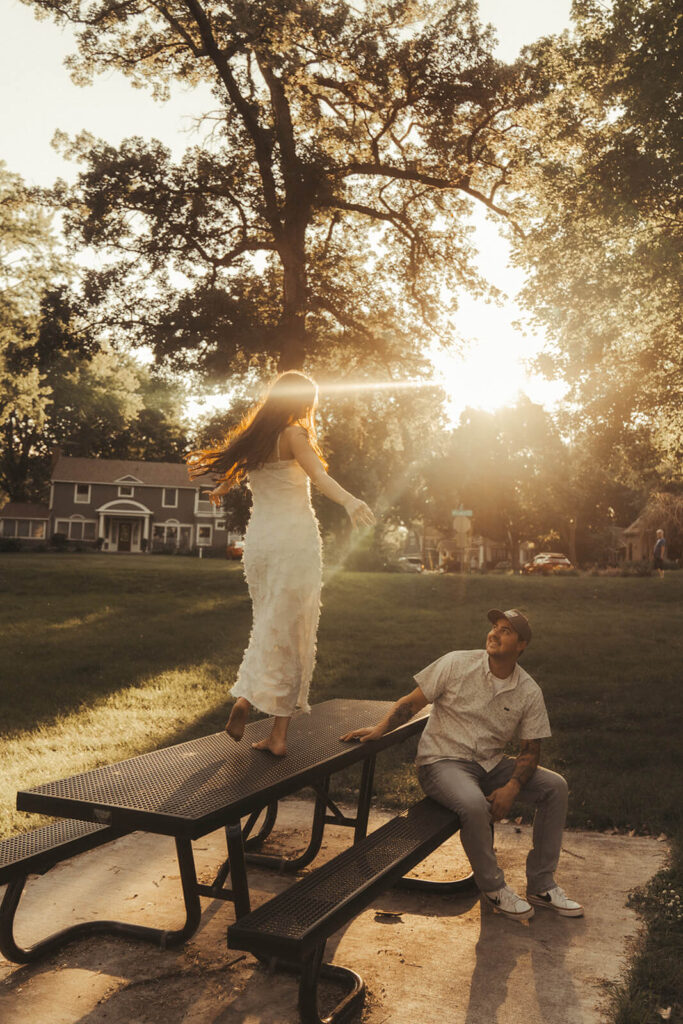 Woman dances on picnic table at golden hour while man looks on.