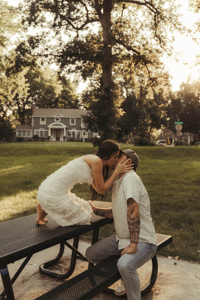 Woman leans down off of picnic table to kiss her fiance.