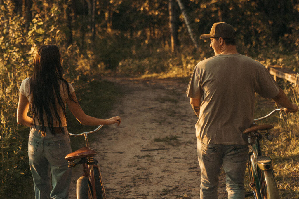 Engaged couple walks down dirt path while wheeling their bikes.