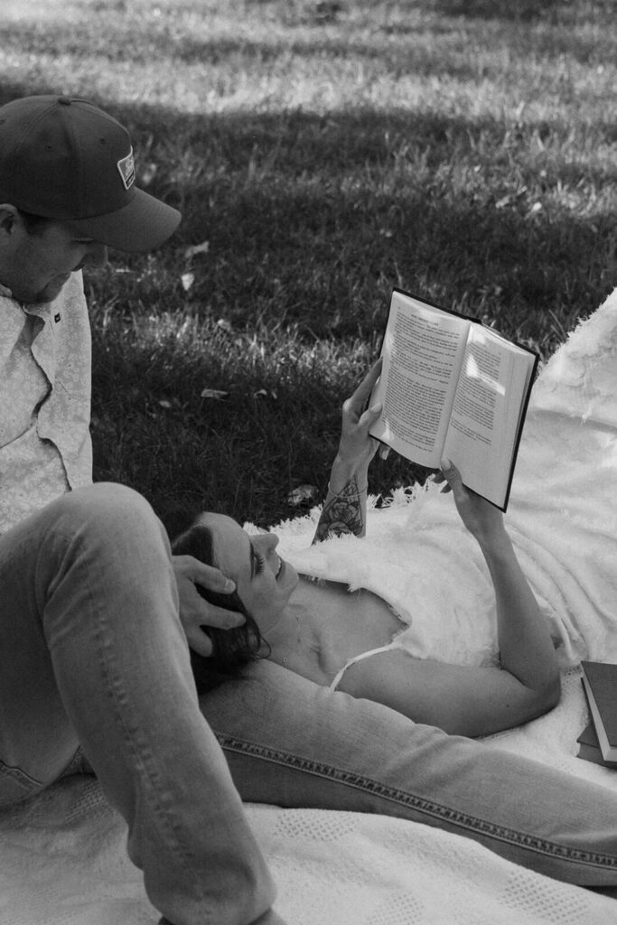 Woman reads book to man as she lays with her head on his leg.