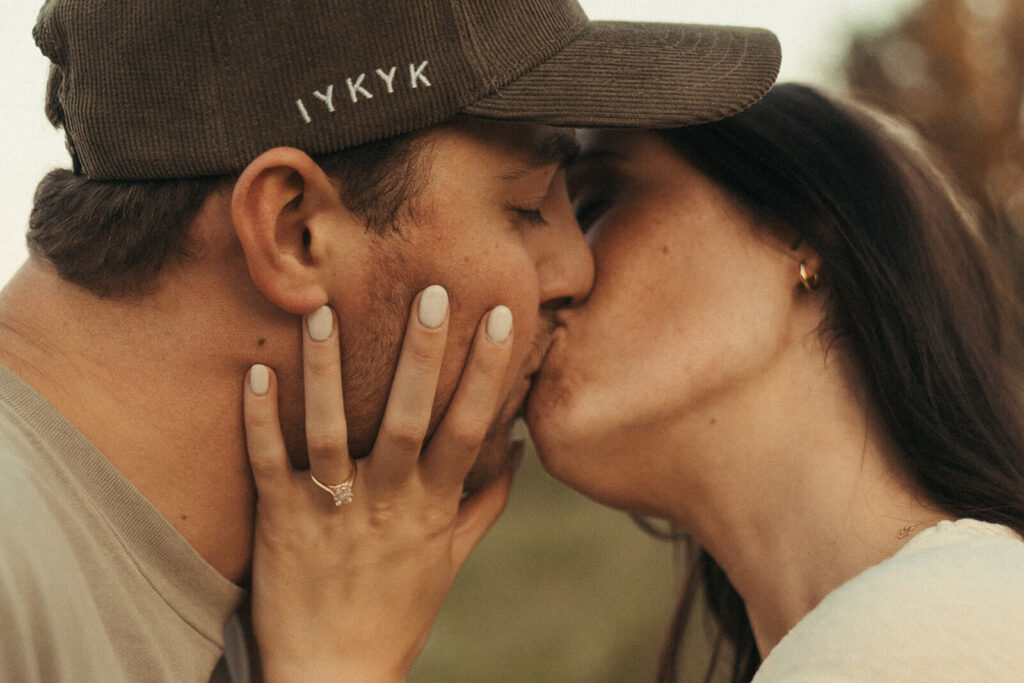 Man wearing ball cap kisses his fiance.