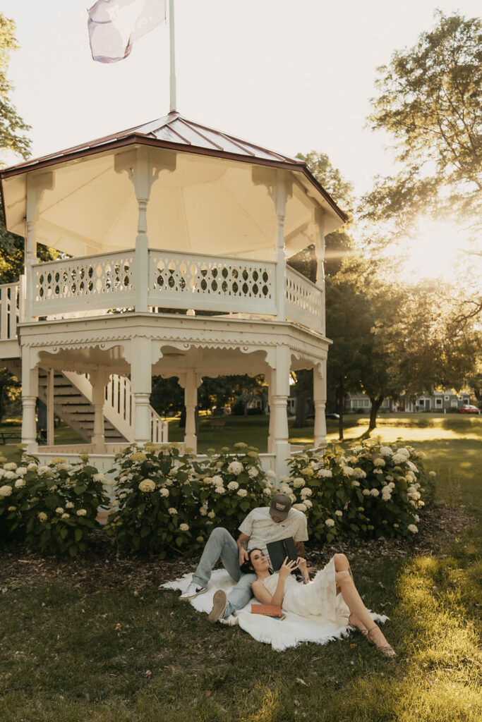 Engaged couple lays in the grass outside gazebo in White Bear Lake, Minnesota as they read a book together.