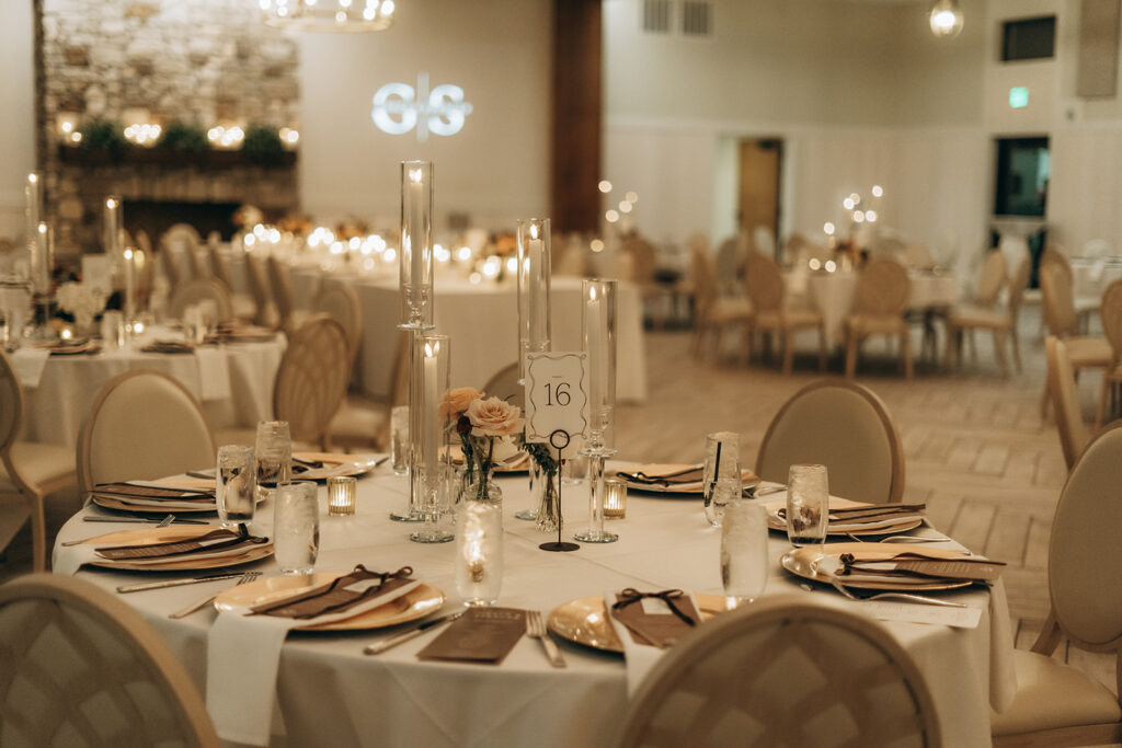 Close-up of a candlelit reception table set with gold chargers, ivory napkins, and a simple floral centerpiece beneath a table number sign, with flickering glass pillar candles adding warmth and glow.