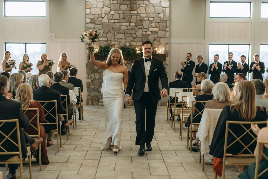 The bride raising her bouquet triumphantly as she and the groom recess down the aisle — just married at their candlelit Chaska wedding.

