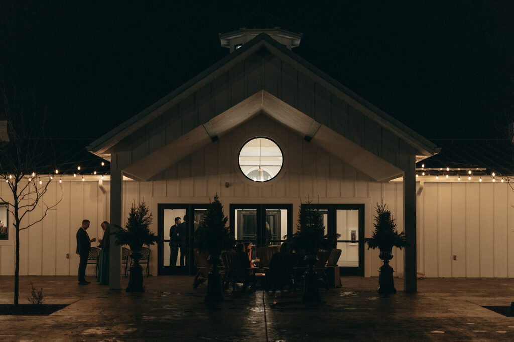 Nighttime exterior of Equestria West, with guests gathered under string lights and around a fire pit, seen through the windows and silhouetted against the white farmhouse-style facade.