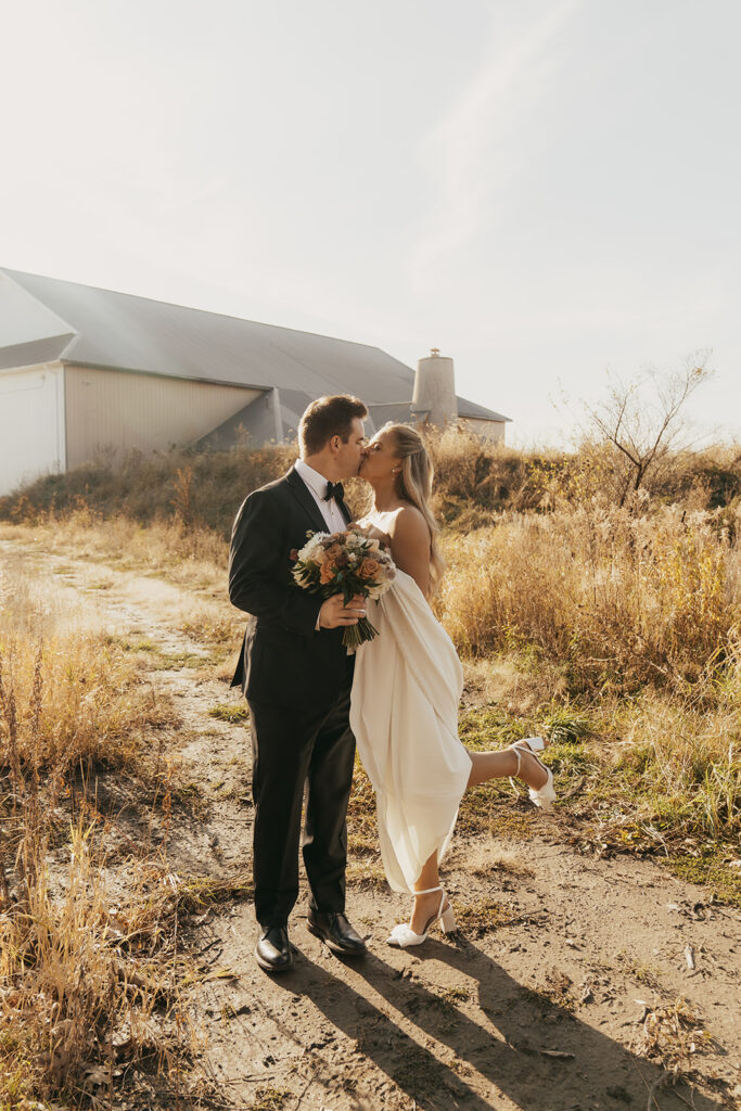 The bride lifts her heel playfully during a kiss near the barn at Equestria West — golden-hour magic from a November wedding in Chaska.