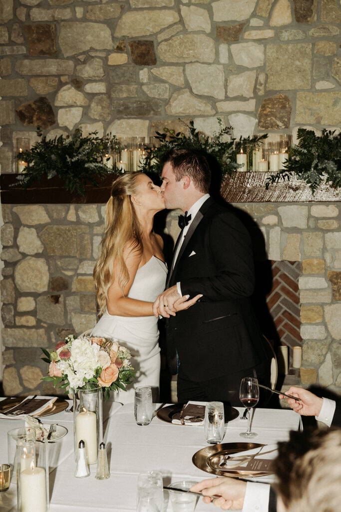The couple shares a kiss at their sweetheart table in front of a candlelit stone fireplace at Equestria West.