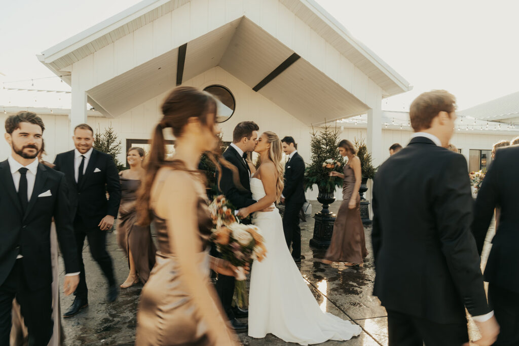 The newlyweds kiss in the courtyard of Equestria West as their wedding party moves around them — golden light meets joyful chaos.
