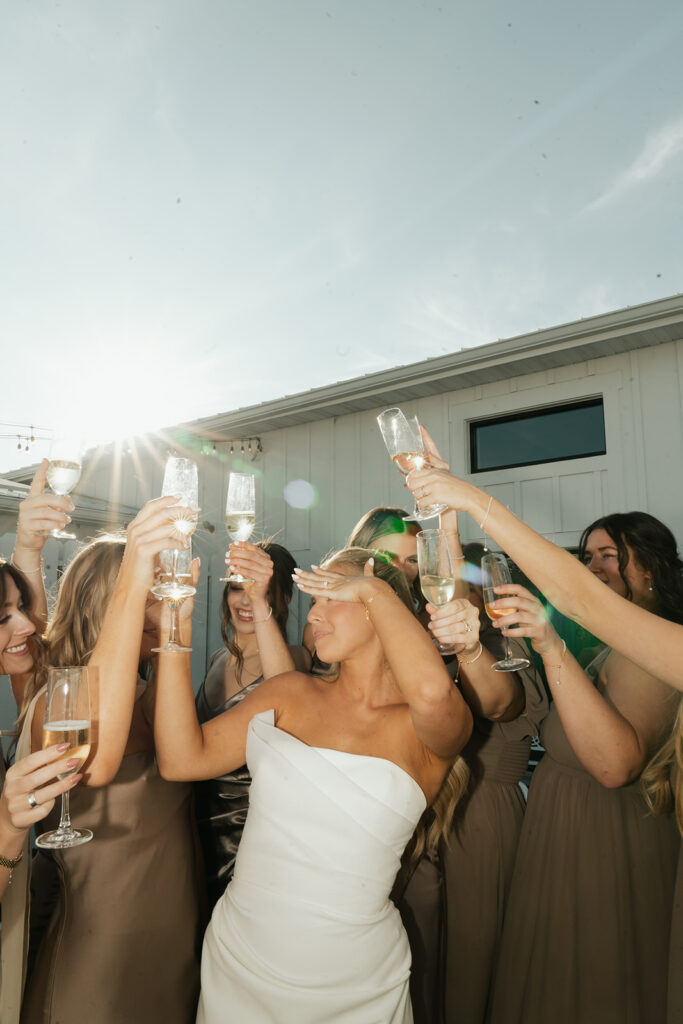 The bride celebrates with bridesmaids at Equestria West, champagne glasses raised against a bright fall sky.