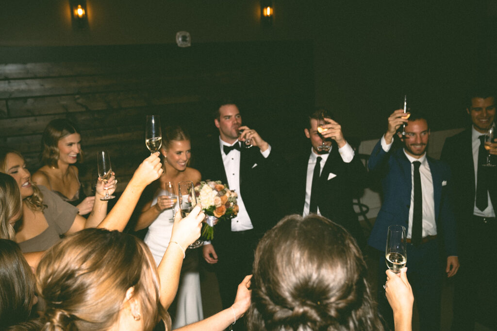 The couple raising champagne glasses with their wedding party in a cozy, warmly lit reception moment in Chaska.


