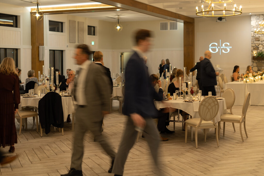 Guests mingling and walking through the softly lit reception space, with tall candles glowing on every table and the couple's initials projected on the back wall.