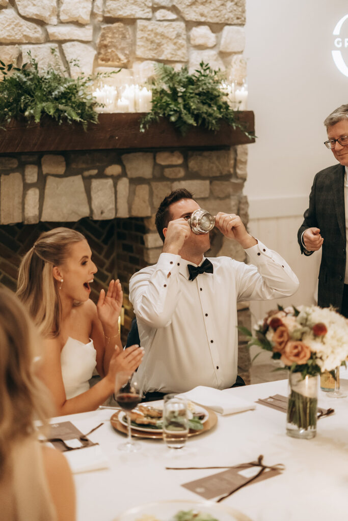Groom cheersing with a pewter mug while guests laugh during the reception at a cozy Chaska venue with stone fireplace and candles.