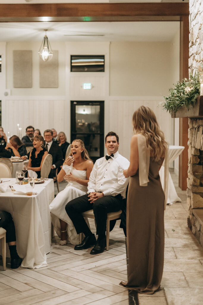 The bride cracking up during a heartfelt speech beside the groom — a cozy, candlelit wedding reception moment at Equestria West.