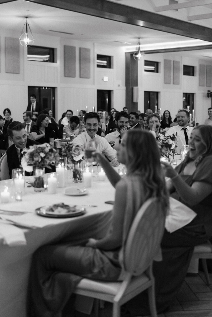 A black-and-white image of guests laughing and toasting at a long head table under soft candlelight.