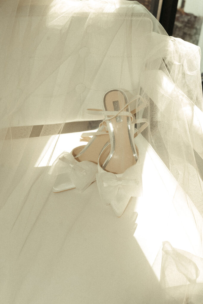 Delicate bridal heels with oversized bows draped in tulle, glowing in soft light before a candlelit wedding ceremony.