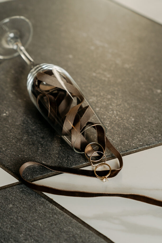 Champagne flute filled with chocolate brown ribbons and wedding rings — styled flatlay from a candlelit Minnesota ceremony.