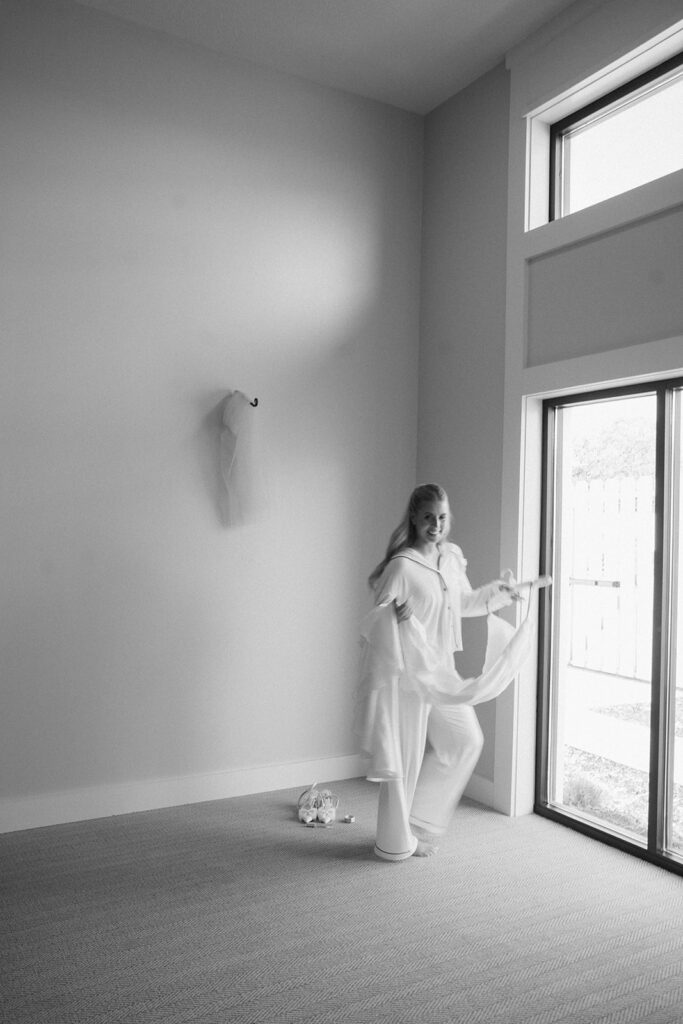 The bride in loungewear playfully holding her wedding dress near tall windows during a quiet Chaska morning.