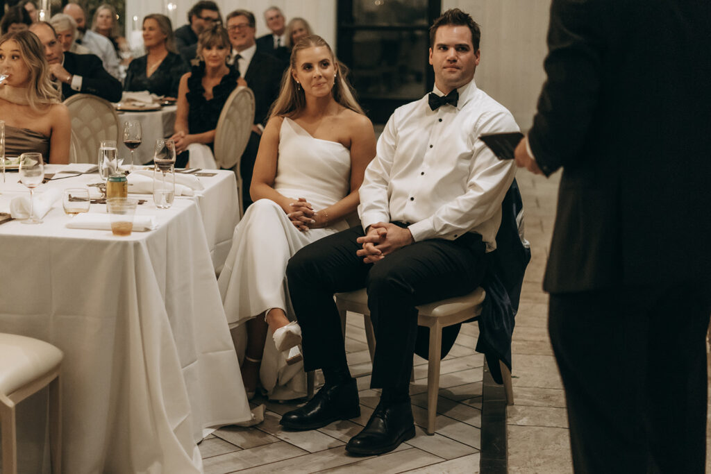 The couple seated side-by-side listening to toasts, framed by tables full of smiling guests, soft candlelight, and a sea of neutral tones and joyful faces.