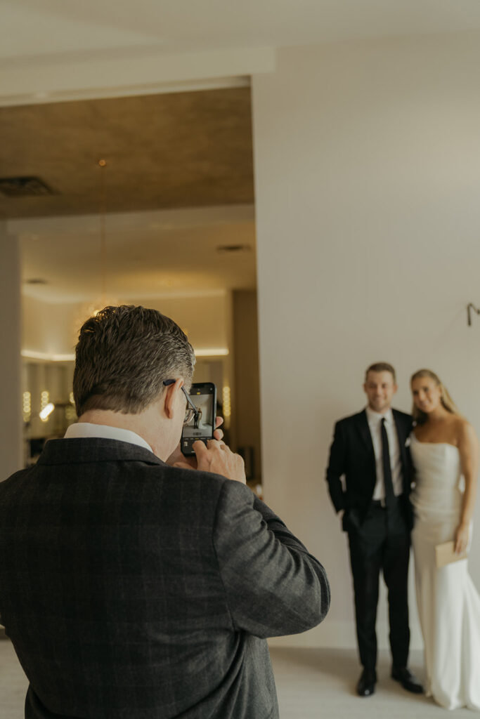 Father of the bride snapping a photo of the bride and her brother — a candid moment before the November wedding begins.
