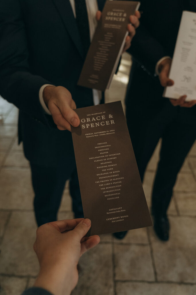 Two groomsmen holding minimalist brown ceremony programs for Grace and Spencer’s November wedding in Chaska, Minnesota.