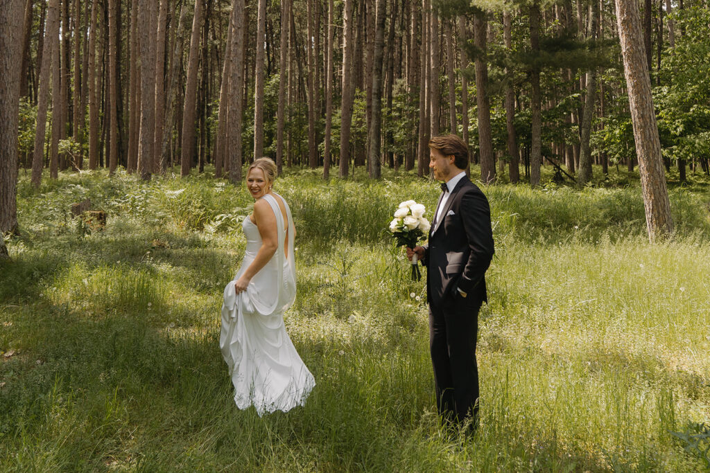 Bride and groom walking through a sun-dappled pine forest at Catalyst by Nature Link, bride smiling back at the camera while holding her dress, groom holding a bouquet.