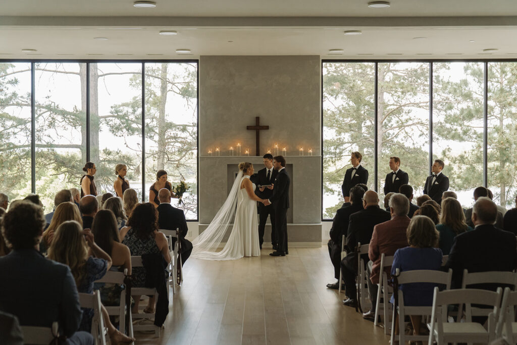 Bride and groom exchanging vows inside a modern chapel at Catalyst by Nature Link, with floor-to-ceiling windows, a wooden cross behind them, and seated guests watching