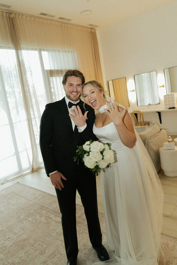 Newlyweds pose with their ring fingers up in the air and expressions of sheer bliss on their faces