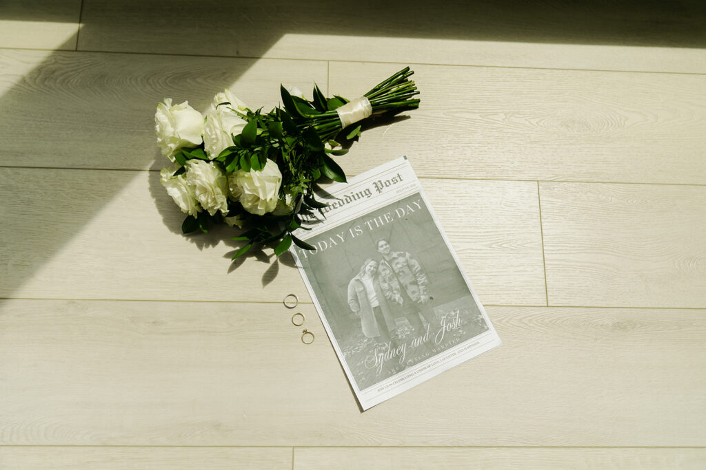 Custom wedding newspaper and bride's bouquet of white roses lay in beam of natural sunlight