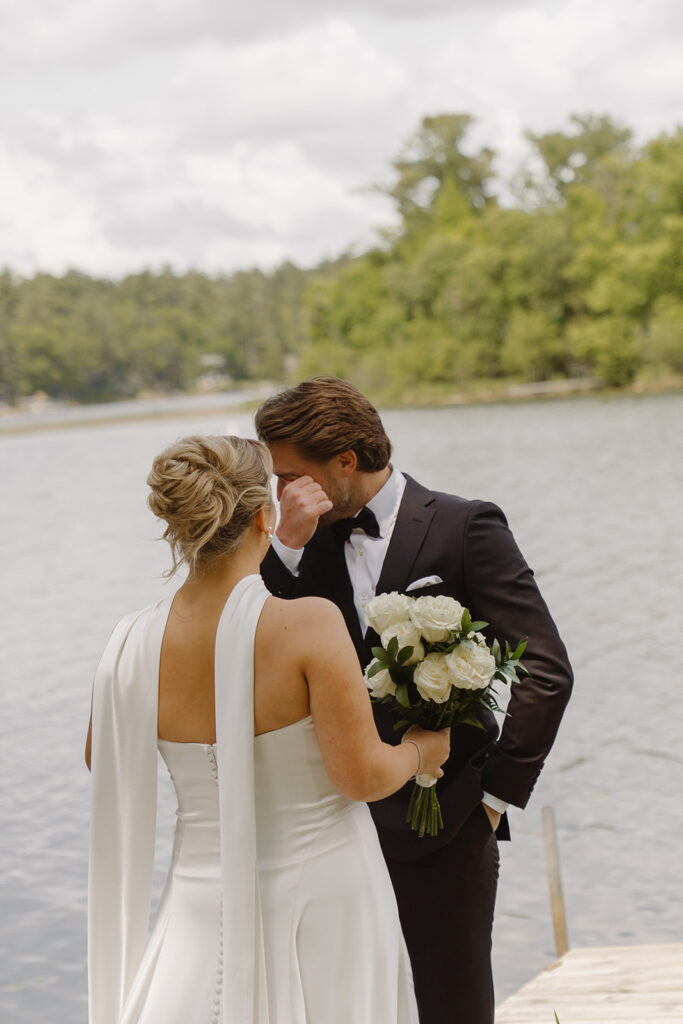 Bride and groom standing on a dock by the lake, bride holding a bouquet of white flowers while groom wipes his tears.