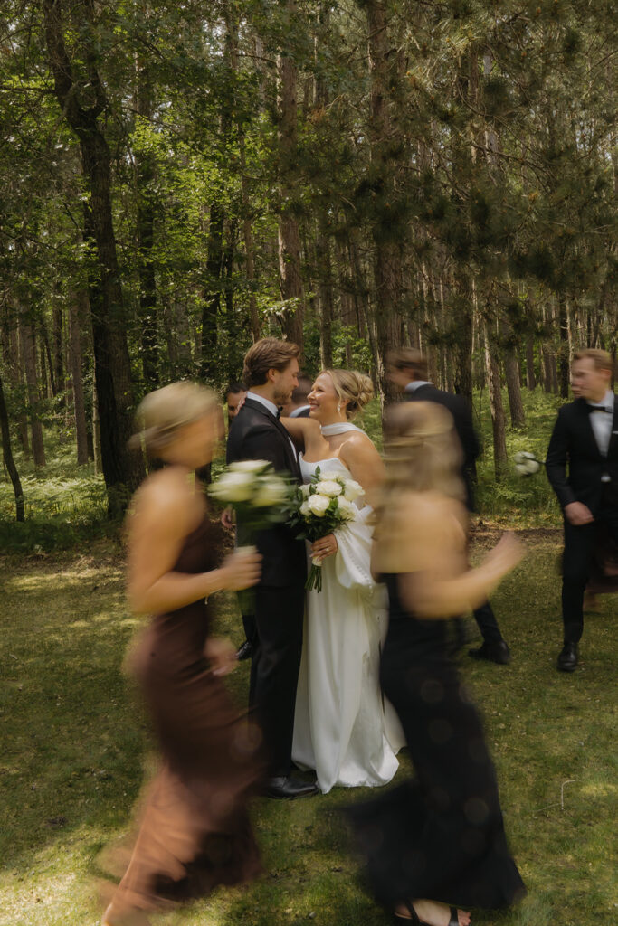 Bridal party moving through a forest, slightly blurred in motion, as bride and groom embrace and smile.