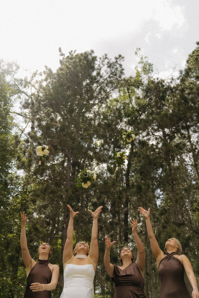 Bride and bridesmaids toss their white rose bouquets into the air with pine tree backdrop
