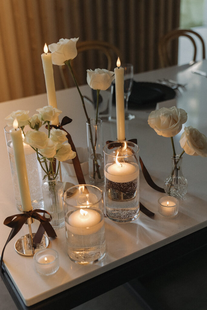Close-up of wedding table with tall taper candles in glass holders and small vases of white roses tied with dark ribbon.