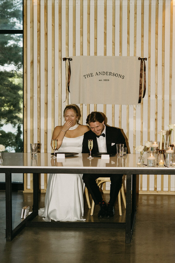 Bride and groom double over in laughter as they listen to speeches