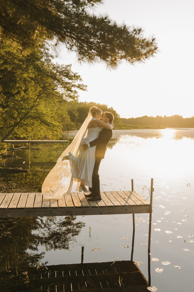 Groom lifts bride into the air as they stand on lakefront dock at golden hour