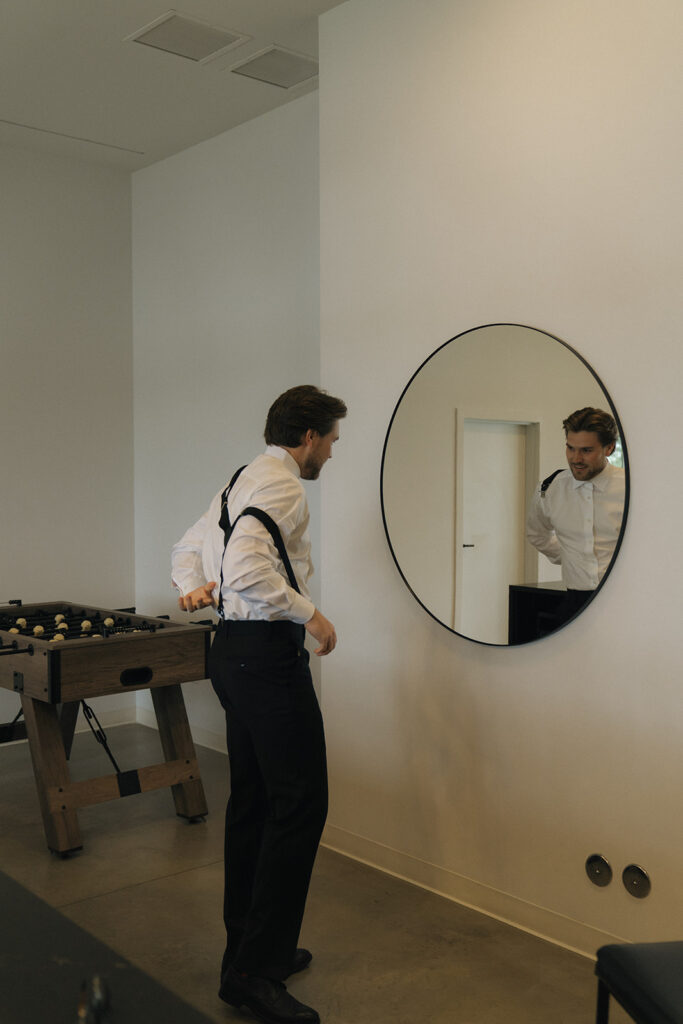 Groom pulls his suspenders up as he looks at his reflection in the mirror