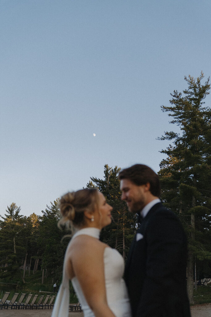 Bride and groom look into each others eyes as moon rises high in the sky during blue hour timeless wedding photos