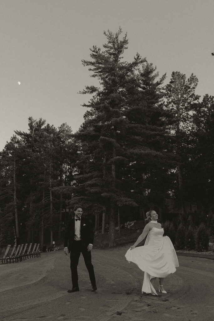 Timeless wedding photos of bride and groom outside under tall Minnesota pine trees at dusk, groom watching bride twirl in her wedding dress, moon visible in sky.