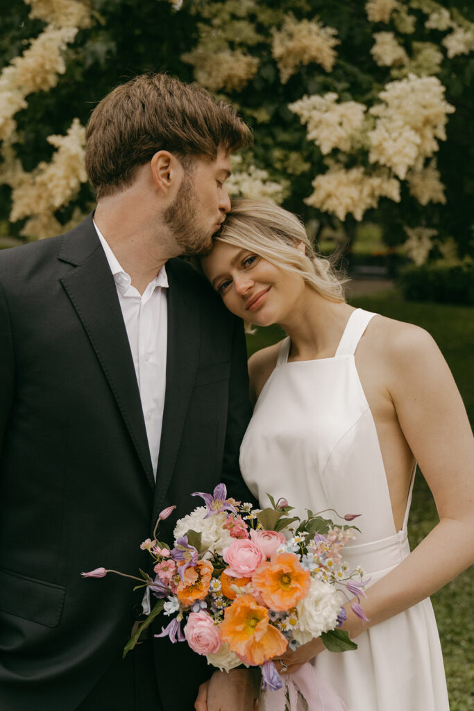Groom kisses bride’s forehead as she smiles, holding bouquet during their Minnesota elopement photos.
