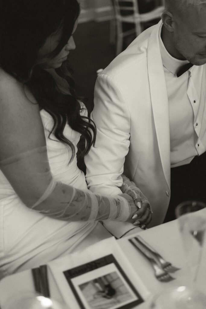 Romantic black-and-white photo of the bride and groom holding hands during their classic wedding dinner.