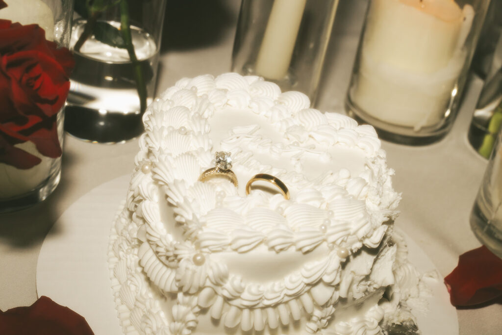 Wedding rings resting atop a vintage heart-shaped cake surrounded by candlelight and rose petals.