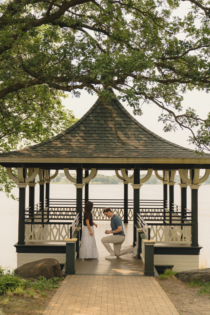 Joey kneeling in the gazebo at Noerenberg Gardens as Bella walks toward him for the proposal, with Lake Minnetonka in the background.