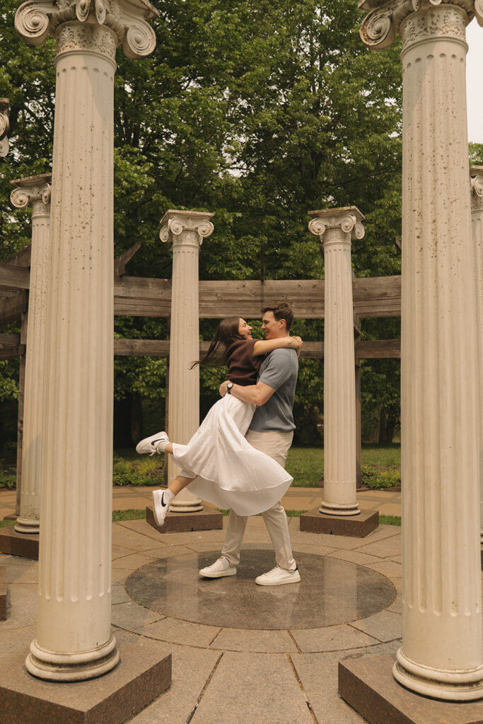 Joey lifting Bella into a spinning hug between the tall white columns at Noerenberg Gardens in Wayzata, MN, celebrating their engagement with playful, romantic wedding proposal photos surrounded by lush summer greenery.