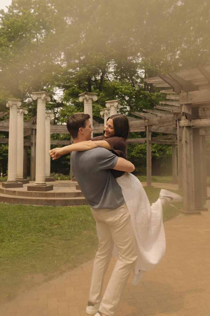 Joey lifting Bella into an embrace near the stone columns, both of them laughing.