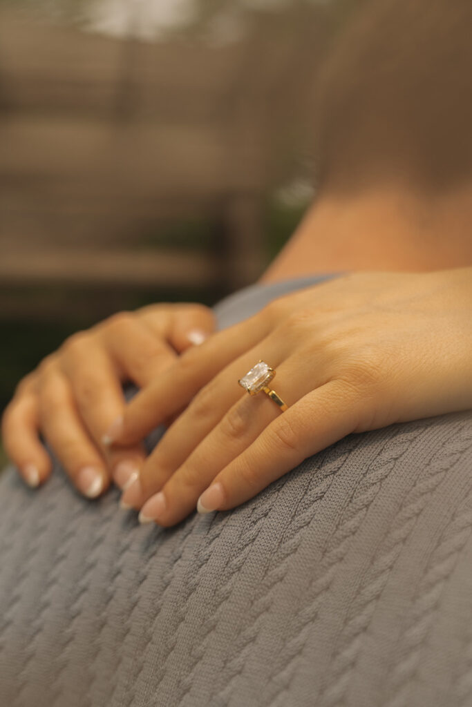 Close-up of an engagement ring on Bella’s hand as she wraps her arms around Joey during their wedding proposal session at Noerenberg Gardens in Wayzata, MN. The gold band and rectangular diamond catch the soft summer light, captured in warm, romantic wedding proposal photography.