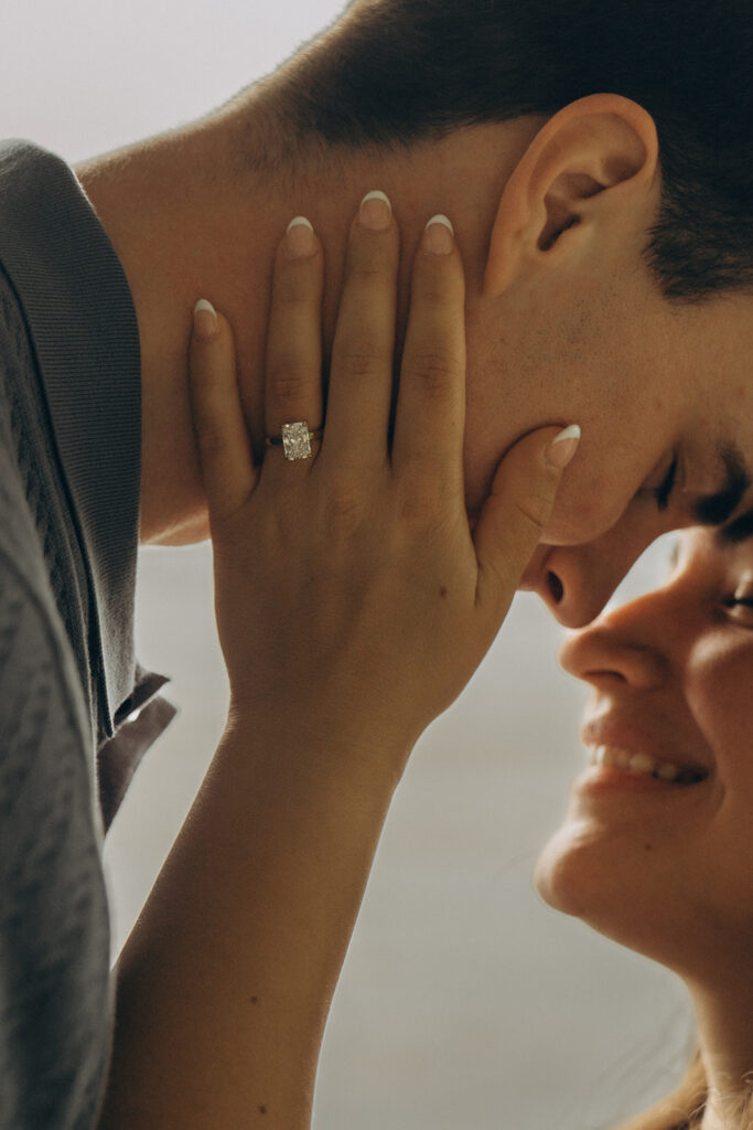 Bella holding Joey’s face with both hands, showing her engagement ring as they lean in close together.