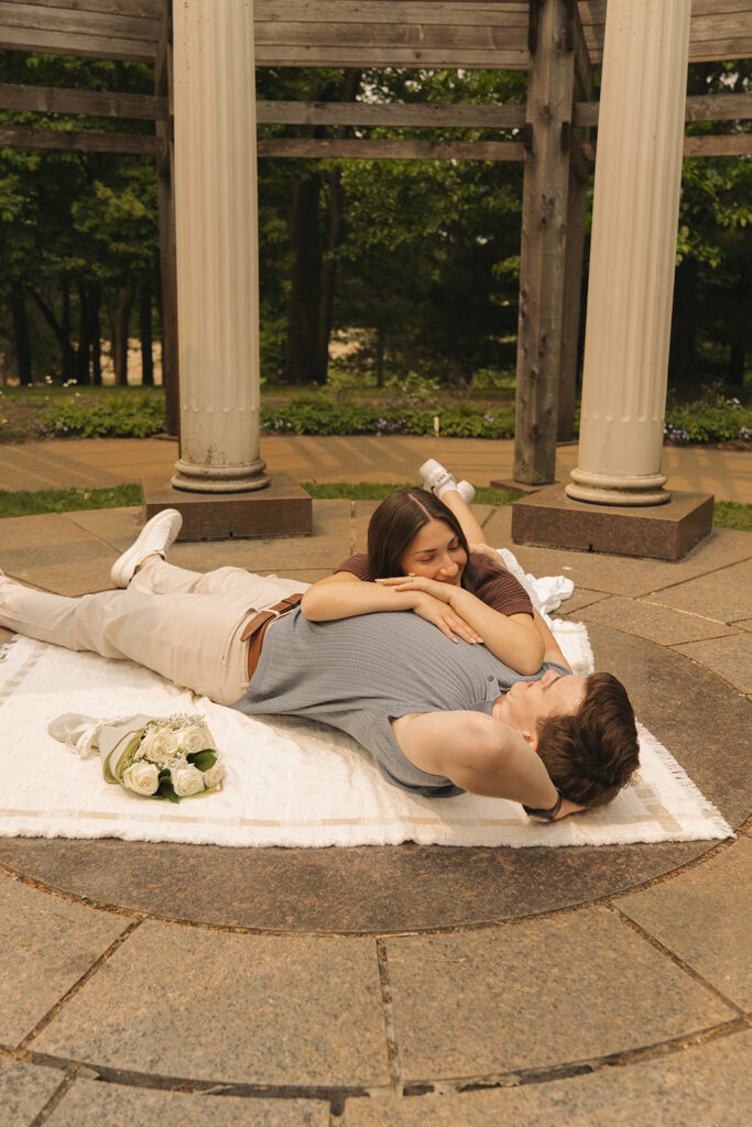 Bella lying on a blanket with her head on Joey’s chest, relaxing together beneath the pergola at Noerenberg Gardens, captured by wedding proposal photographer.