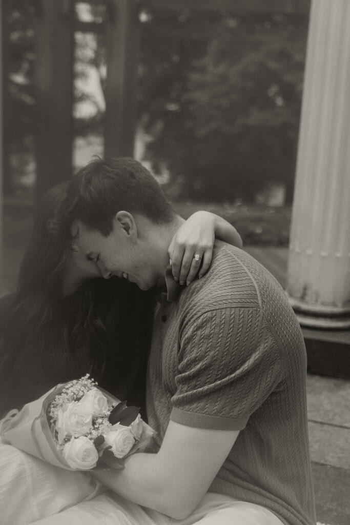 Joey and Bella sitting close together after the proposal, laughing as she leans into his shoulder and holds her bouquet.
