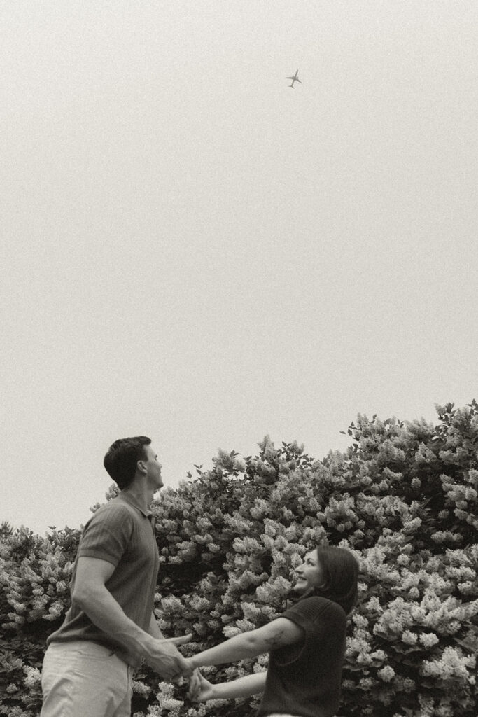 Joey and Bella standing beneath blooming bushes, holding hands and looking up toward a small plane overhead.