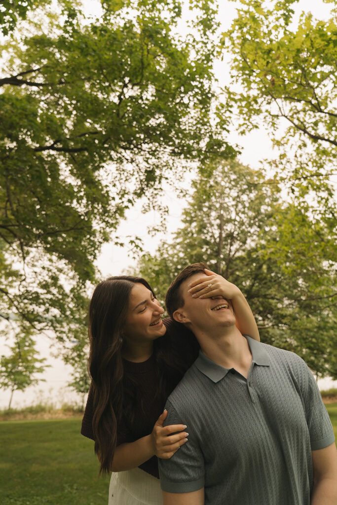 Bella wrapping her hands around Joey's eyes from behind as they laugh under the trees during their engagement photos in Wayzata, MN.